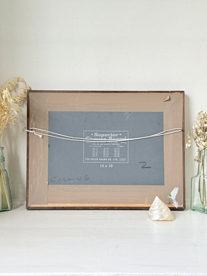 A vintage wooden picture frame with a clear glass front, held together by a white cord. The frame is set on a white surface against a white wall, flanked by dried grass in glass bottles and a small seashell.
