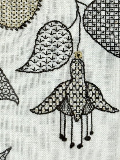 Framed circular hand embroidery of birds and fuchsia flowers in black and grey on white linen, set in gold frame, white surface background.