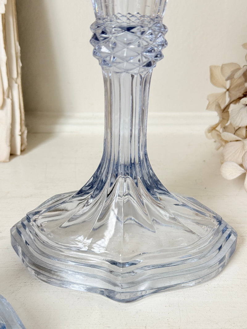 A clear cut-glass candlestick holder with a flared base and textured neck, shown on a light tabletop with soft neutral background decor.