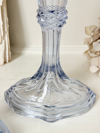 A clear cut-glass candlestick holder with a flared base and textured neck, shown on a light tabletop with soft neutral background decor.