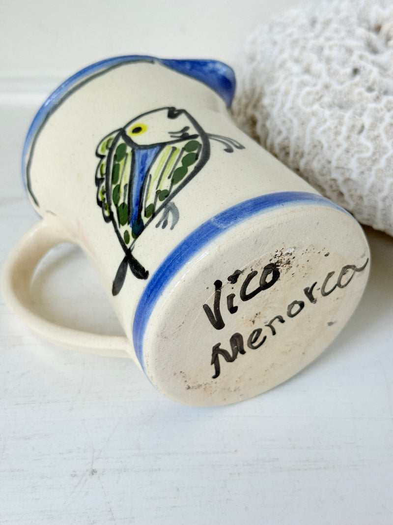 Vintage studio pottery jug with hand painted blue and green fish design on cream clay, photographed on a white surface.