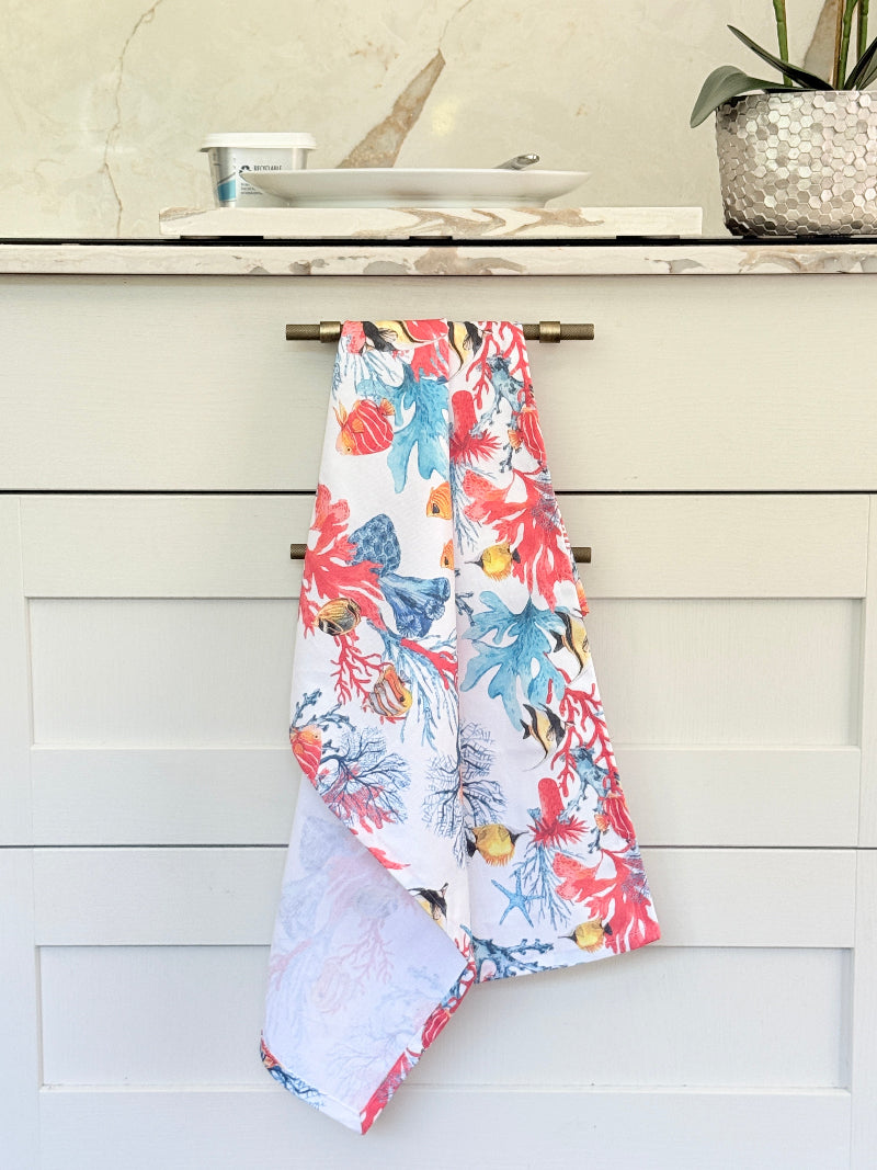 Coral reef print cotton tea towel in crisp white with colourful coral and tropical fish motifs, photographed indoors