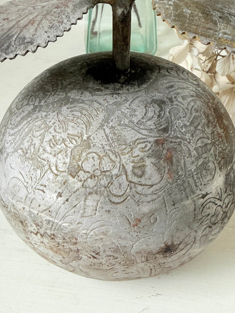 An antique silver metal apple-shaped vase with intricate embossed floral patterns, featuring a dark metal stem and leaf-like top details, set against a light background with dried flowers.