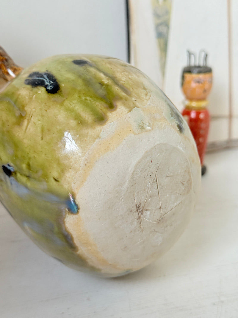 A close-up of a handmade ceramic vessel, showing the glossy glaze with green, beige, and blue variations on one side and a raw, unglazed beige clay base on the other. A small red and orange wooden doll is visible in the blurred background.