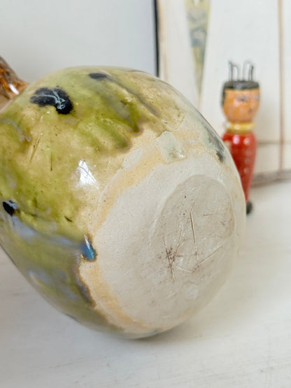 A close-up of a handmade ceramic vessel, showing the glossy glaze with green, beige, and blue variations on one side and a raw, unglazed beige clay base on the other. A small red and orange wooden doll is visible in the blurred background.