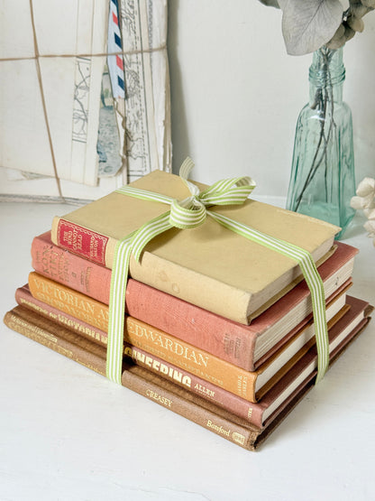 A stack of vintage hardcover books tied with a light green and white striped ribbon, set on a white surface with a rustic background featuring a blue glass bottle and framed artwork.