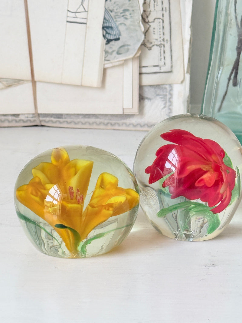 Pair of orange and yellow flower glass paperweights with green leaves, photographed on a white surface