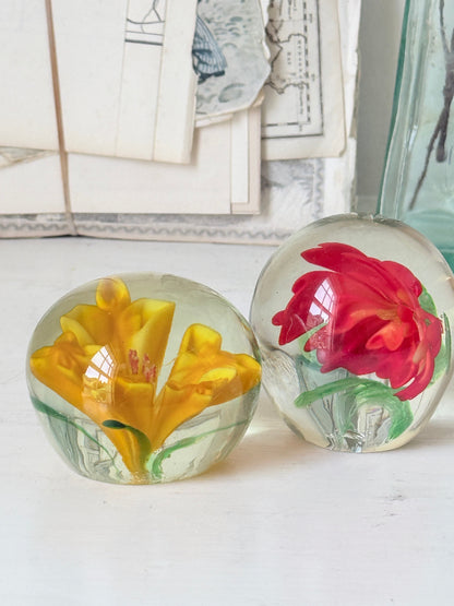 Pair of orange and yellow flower glass paperweights with green leaves, photographed on a white surface