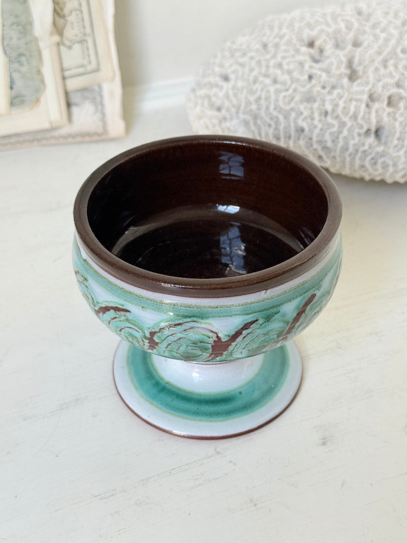 Hand thrown studio pottery footed bowl with green and brown swirling decoration photographed on a white surface