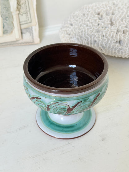 Hand thrown studio pottery footed bowl with green and brown swirling decoration photographed on a white surface