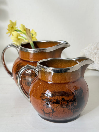 Pair of antique Ridgways stoneware creamer jugs photographed on a white surface in amber treacle glaze with black coaching scenes and silver lustre rims