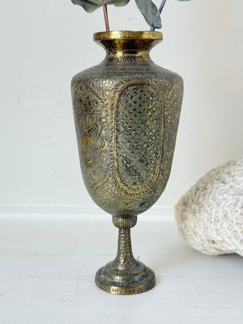 Indian Benares brass chalice vase with figural panels and pedestal base, photographed on a white surface