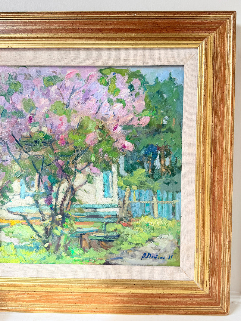 A framed impressionist oil painting depicting a blooming pink tree in front of a white house with blue shutters, enclosed in a gold frame with a cream mat.