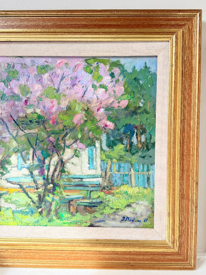 A framed impressionist oil painting depicting a blooming pink tree in front of a white house with blue shutters, enclosed in a gold frame with a cream mat.