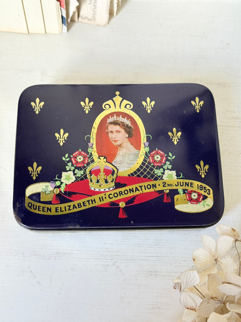 A rectangular dark blue tin with rounded corners, featuring a portrait of Queen Elizabeth II, a crown, fleur-de-lis motifs, and a banner reading &
