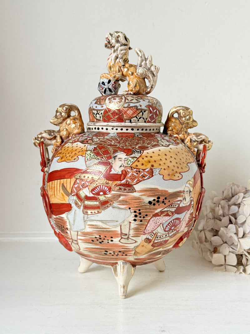 An ornate ceramic lidded jar with three legs, featuring hand-painted figures and patterns in red, gold, and white, with decorative animal figures on the lid and side handles.