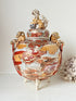 An ornate ceramic lidded jar with three legs, featuring hand-painted figures and patterns in red, gold, and white, with decorative animal figures on the lid and side handles.
