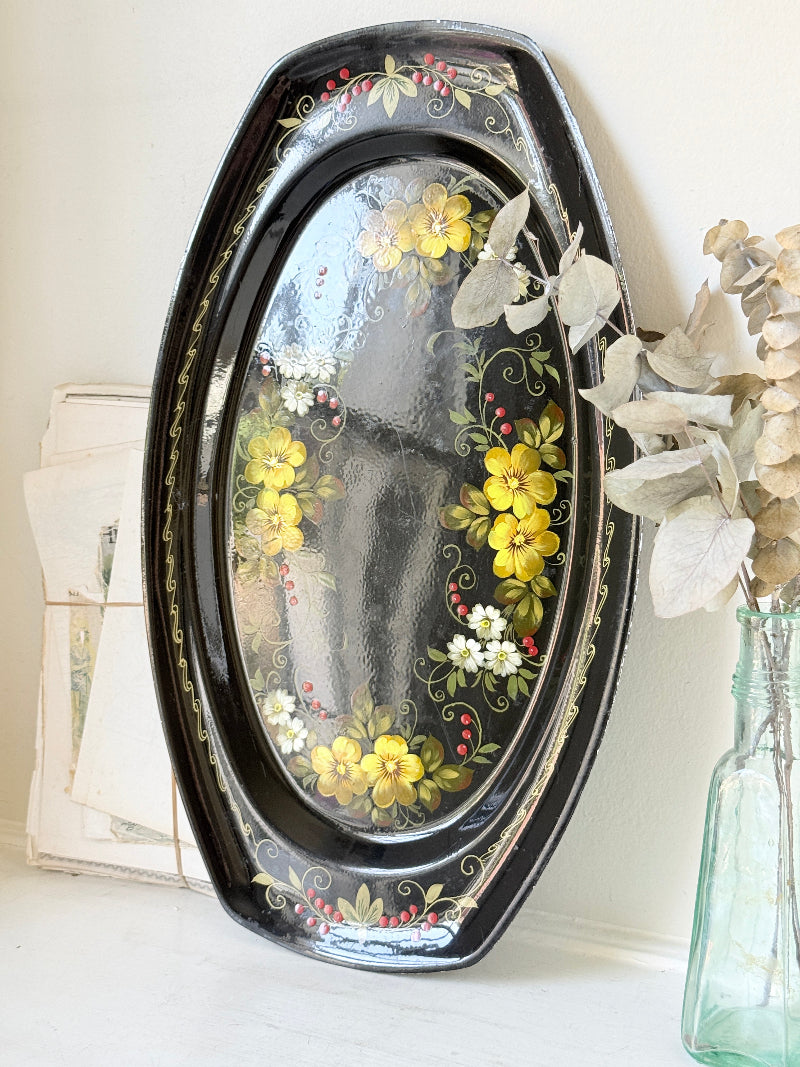 An oval black decorative tray with hand-painted yellow flowers, green leaves, and small white blossoms, leaning against a light wall next to a glass bottle with dried flowers.