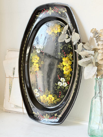 An oval black decorative tray with hand-painted yellow flowers, green leaves, and small white blossoms, leaning against a light wall next to a glass bottle with dried flowers.