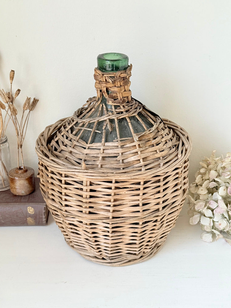antique French demi john olive oil jar in wicker basket with green glass bottle and painted metal label, displayed on white surface