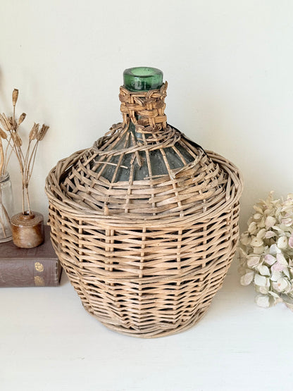 antique French demi john olive oil jar in wicker basket with green glass bottle and painted metal label, displayed on white surface