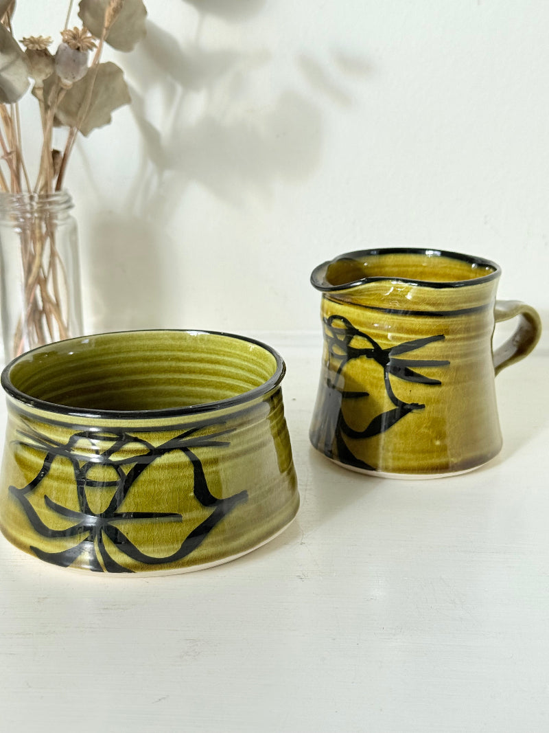 Studio pottery sugar bowl and milk jug in olive green glaze with black brushstroke decoration, photographed on a white surface.