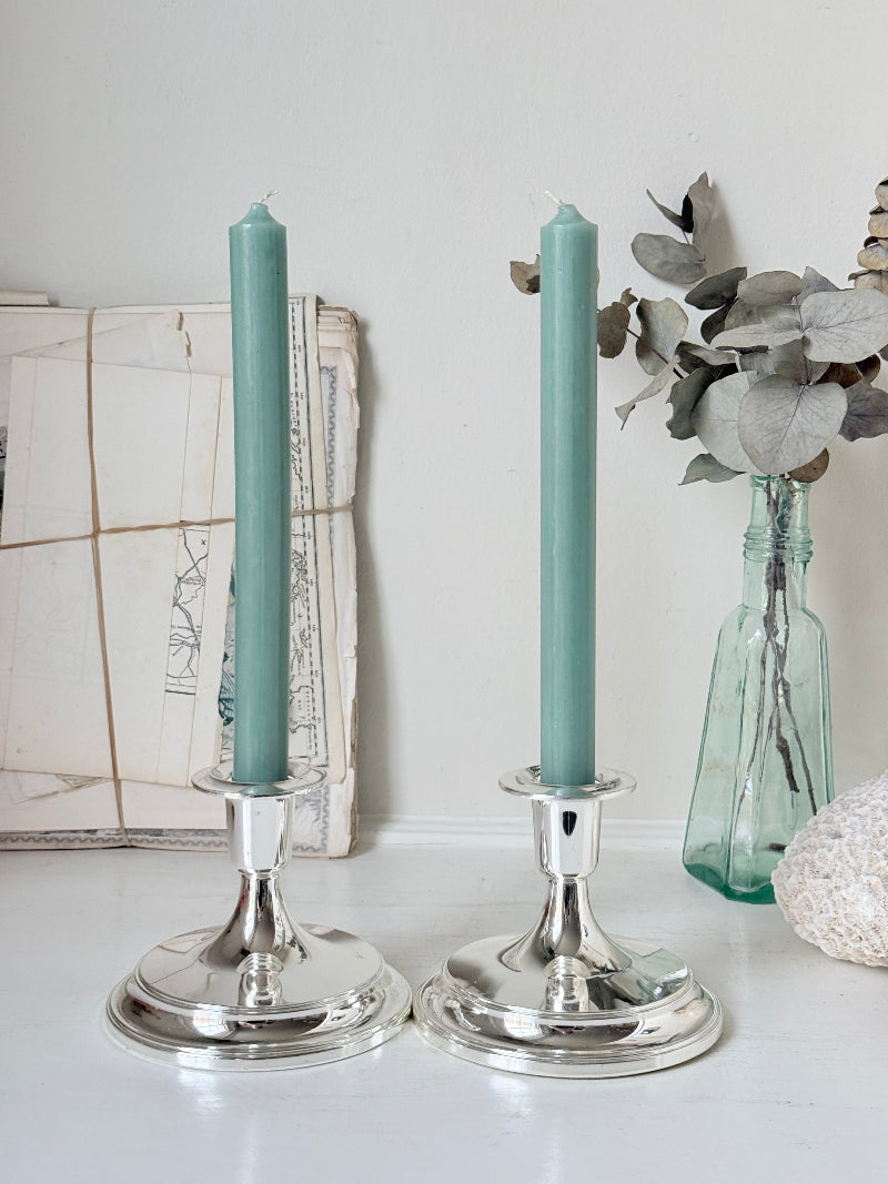 Pair of silver tone candlesticks with wide round stepped bases holding green candles, photographed on a white surface