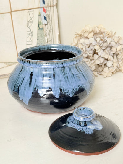A ceramic lidded jar with a glossy black and light blue glaze, shown with its lid placed in front on a light surface. The background includes a decorative dried flower and a framed print.