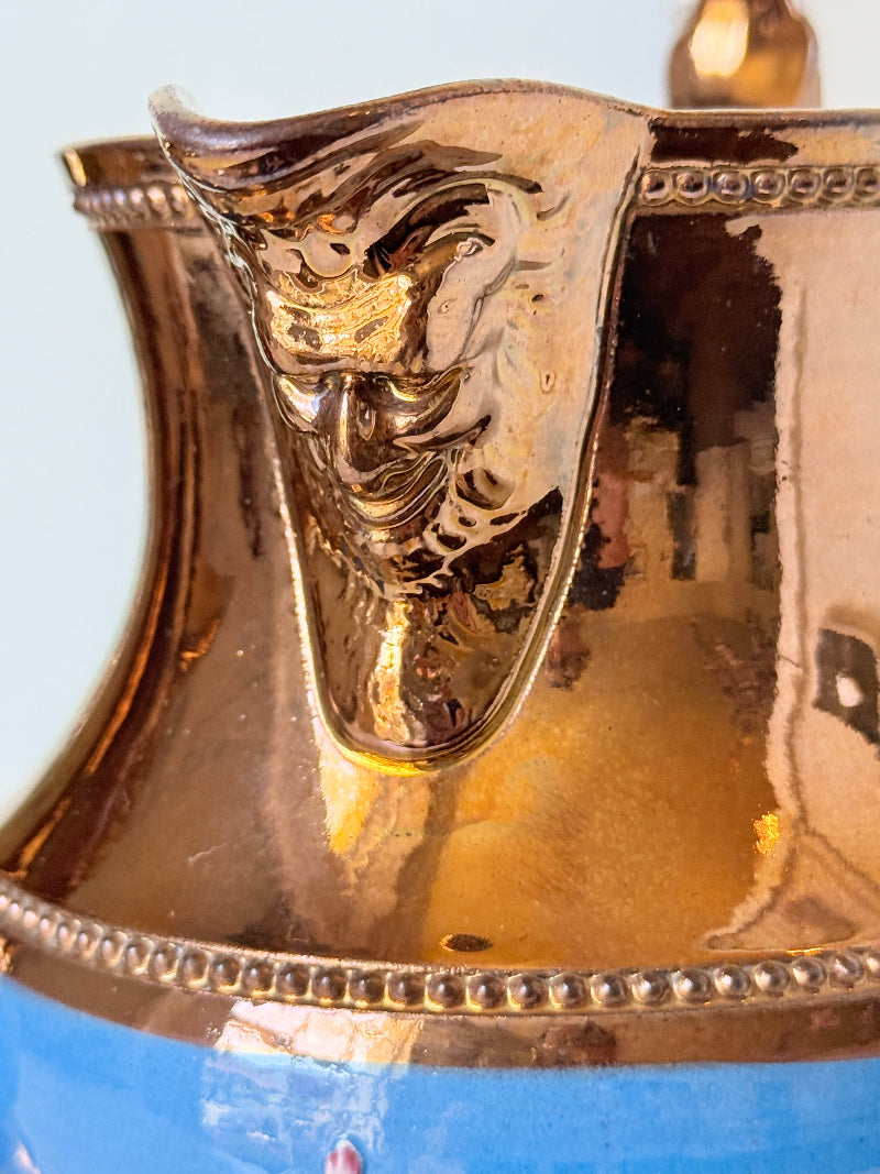 Close-up of a metallic gold pitcher with a sculpted lion head spout and a blue lower section, reflecting a real-world background.