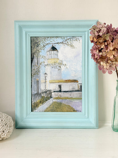 A framed watercolor painting of a lighthouse, featuring a light blue painted frame, displayed on a tabletop next to a vase of dried flowers.