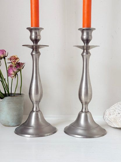 Pair of vintage pewter effect baluster candlesticks with orange taper candles styled on a white surface