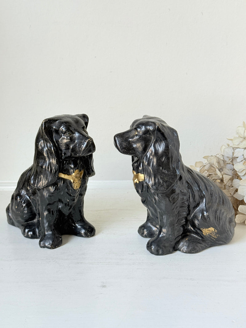 Two black ceramic dog figurines with gold accents, resembling Cocker Spaniel dogs, displayed on a white surface against a light background.