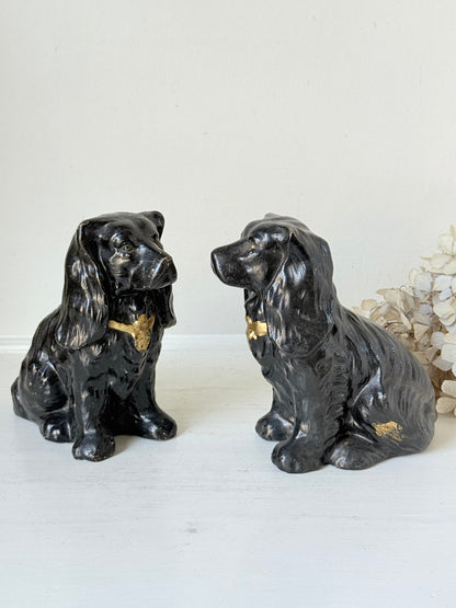 Two black ceramic dog figurines with gold accents, resembling Cocker Spaniel dogs, displayed on a white surface against a light background.