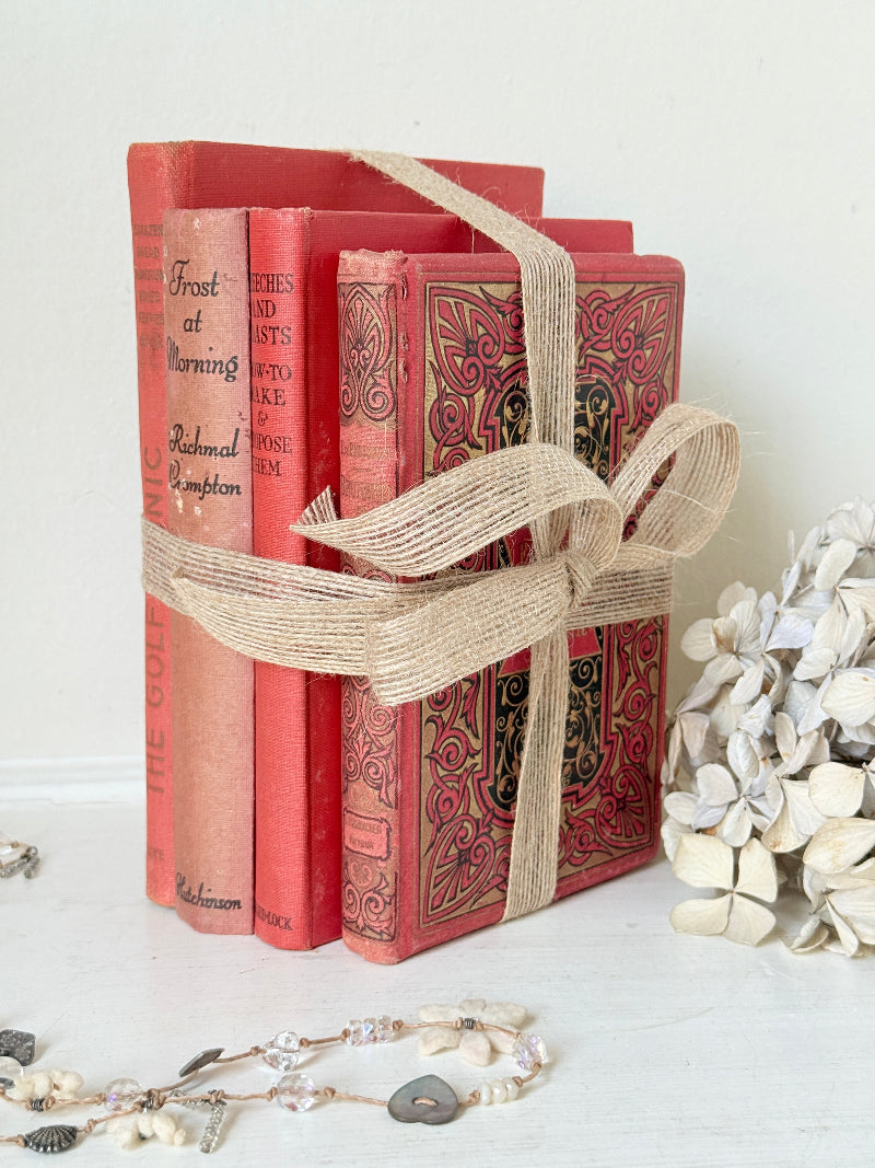 Three vintage-style red books tied together with a beige burlap ribbon, displayed on a white surface with dried flowers and jewelry props.