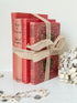 Three vintage-style red books tied together with a beige burlap ribbon, displayed on a white surface with dried flowers and jewelry props.