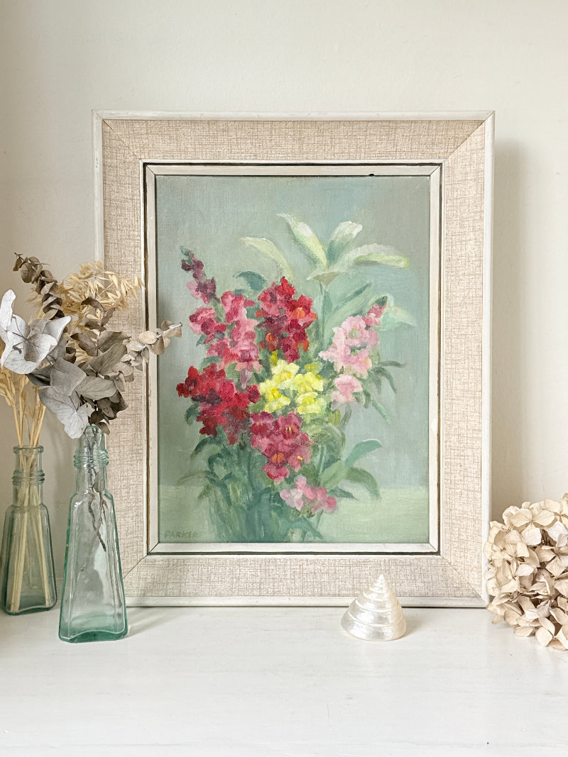A framed floral painting featuring vibrant pink, red, and yellow flowers with green leaves on a soft blue-green background, housed in a textured beige linen-style frame. The frame is styled on a white surface next to glass vases with dried flowers and a small shell.