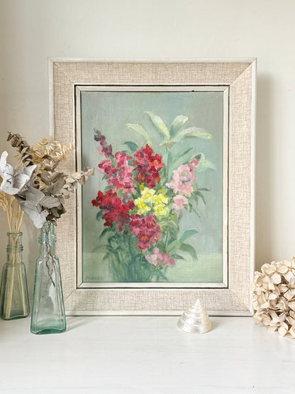 A framed floral painting featuring vibrant pink, red, and yellow flowers with green leaves on a soft blue-green background, housed in a textured beige linen-style frame. The frame is styled on a white surface next to glass vases with dried flowers and a small shell.