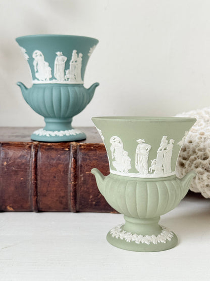 Pair of vintage Wedgwood Jasperware pedestal urns in sage green and pale blue, decorated with white neoclassical figures, styled on an antique leather book.
