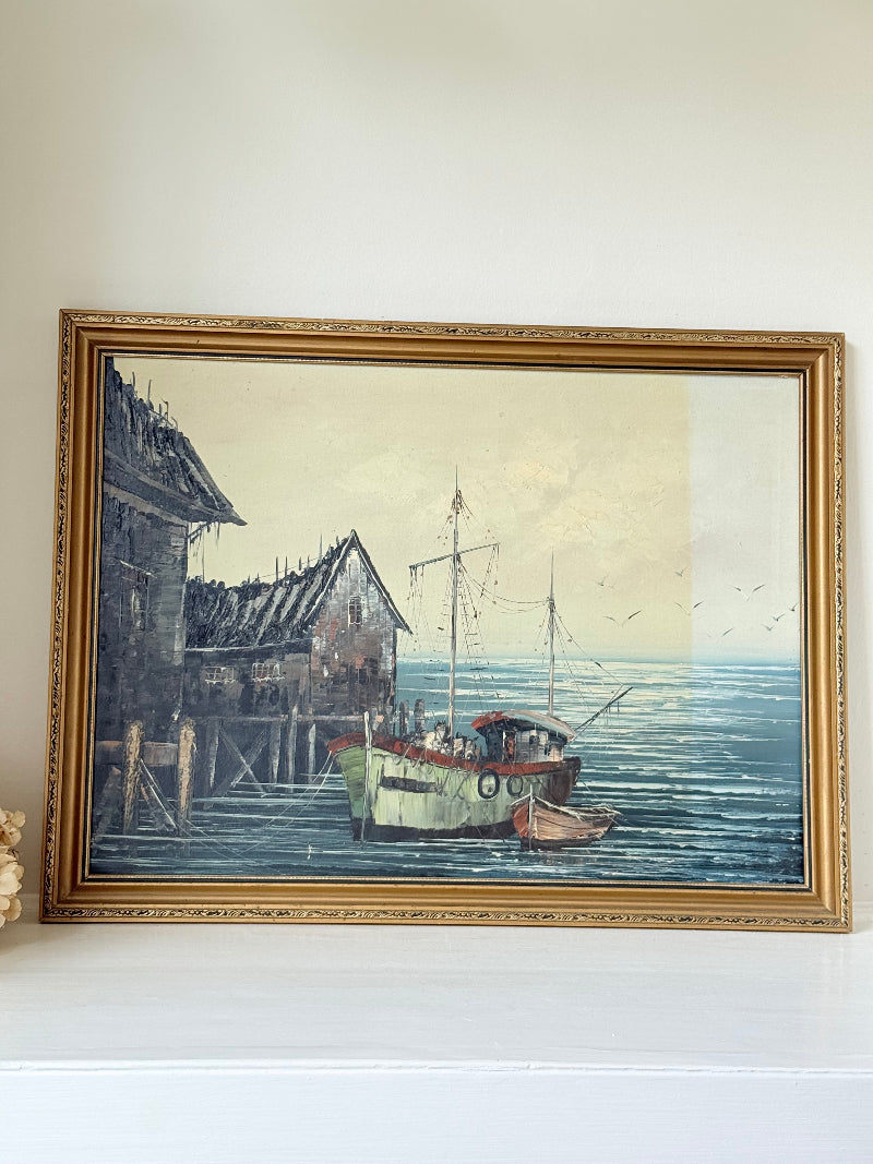 Vintage harbour scene oil painting of fishing boat and wharf buildings in gilt frame, textured brushwork, on white surface.