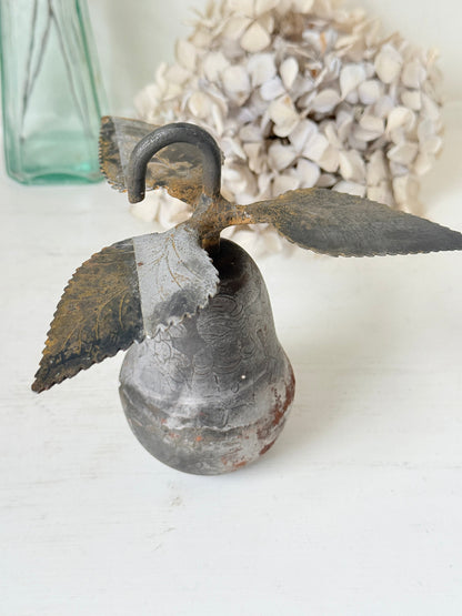 A rustic metal bird-shaped bell with a dark gray and weathered brown patina, displayed on a white surface with a blurred glass vase and dried flowers in the background.
