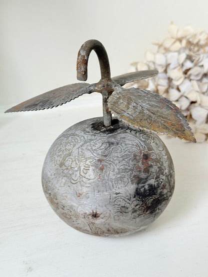 A rustic metal apple-shaped decorative object with engraved details, a curved hook handle, and leaf-like side extensions, photographed on a light surface with a soft blurred background.