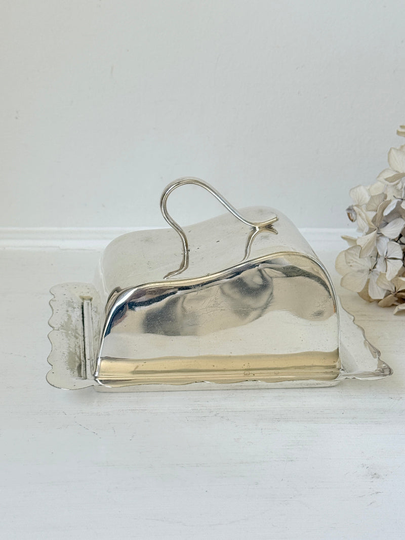 A vintage polished silver metal butter dish with a hinged lid and scalloped sides, resting on a white wooden surface against a white wall.
