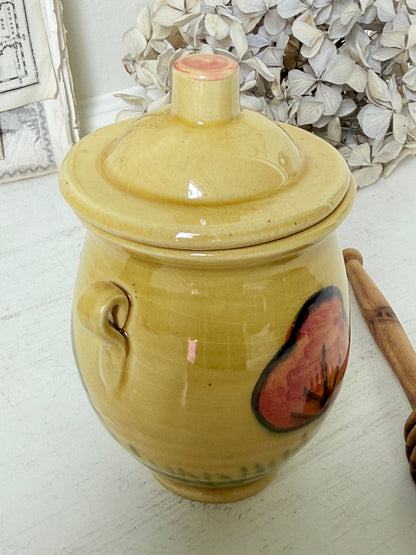 A handmade yellow ceramic lidded jar with a side handle and a painted red and black butterfly design on the front, set on a white surface with dried flowers in the background.