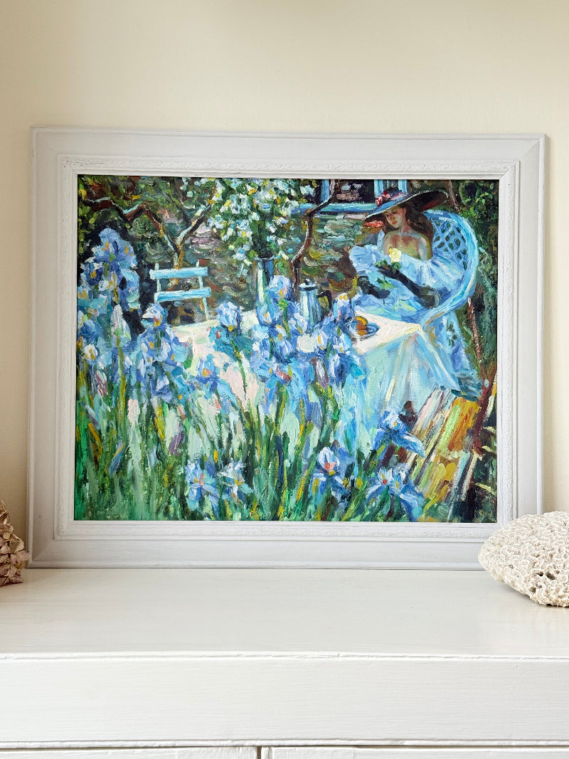 Framed impressionist-style oil painting of blue irises and garden scene, photographed on a white surface