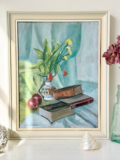 A framed still life painting displayed on a tabletop. The artwork depicts a vase of yellow flowers and green leaves beside stacked books and red apples, set against a soft blue-green draped background.