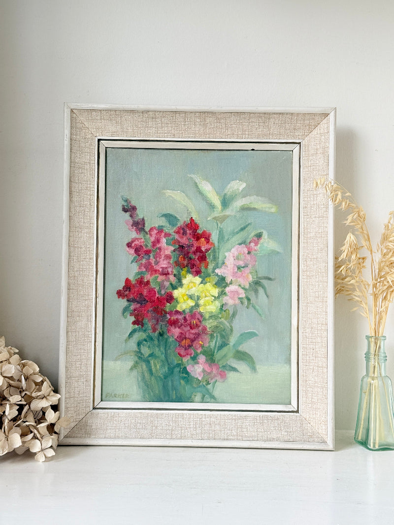 A framed floral painting featuring vibrant pink, red, and yellow flowers with green leaves on a soft blue-green background, housed in a light beige textured frame and styled on a white surface with dried flowers.