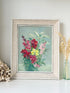 A framed floral painting featuring vibrant pink, red, and yellow flowers with green leaves on a soft blue-green background, housed in a light beige textured frame and styled on a white surface with dried flowers.