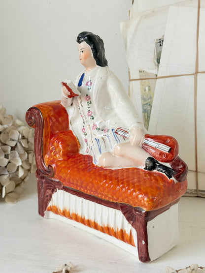 A vintage ceramic figurine of a man reclining on an orange-red chaise lounge, set against a softly blurred background.