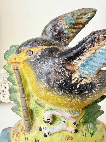 A close-up of a vintage hand-painted ceramic bird figurine, likely a robin, perched on a green leafy branch and holding a bamboo flute. The bird features a distressed finish with black, yellow, blue, and green glazes.