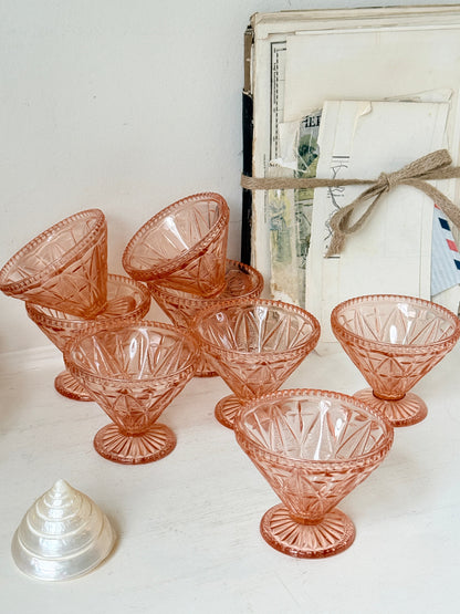 A set of vintage pink cut-glass cocktail glasses arranged on a white surface, with a small silver seashell in the foreground and a rustic framed print tied with twine in the background.